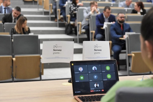Na ekranie laptopa widoczne są kafelki osób, które będą zdawały egzamin on-line, w tle siedzą uczestnicy studiów podyplomowych