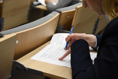 Siedząca funkcjonariuszka pochylona nad arkuszem egzaminacyjnym, w ręku trzyma długopis