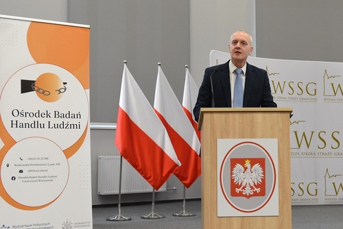 Po egzaminie gen. bryg. SG dr Piotr Boćko otwiera konferencję. Na mównicy widać godło państwowe, natomiast w tle widać banery i biało-czerwone flagi